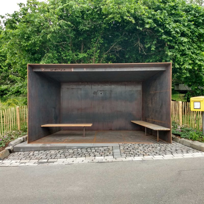 Haltestelle aus Stahl mit Holzbänken, davor abschüssige Straße