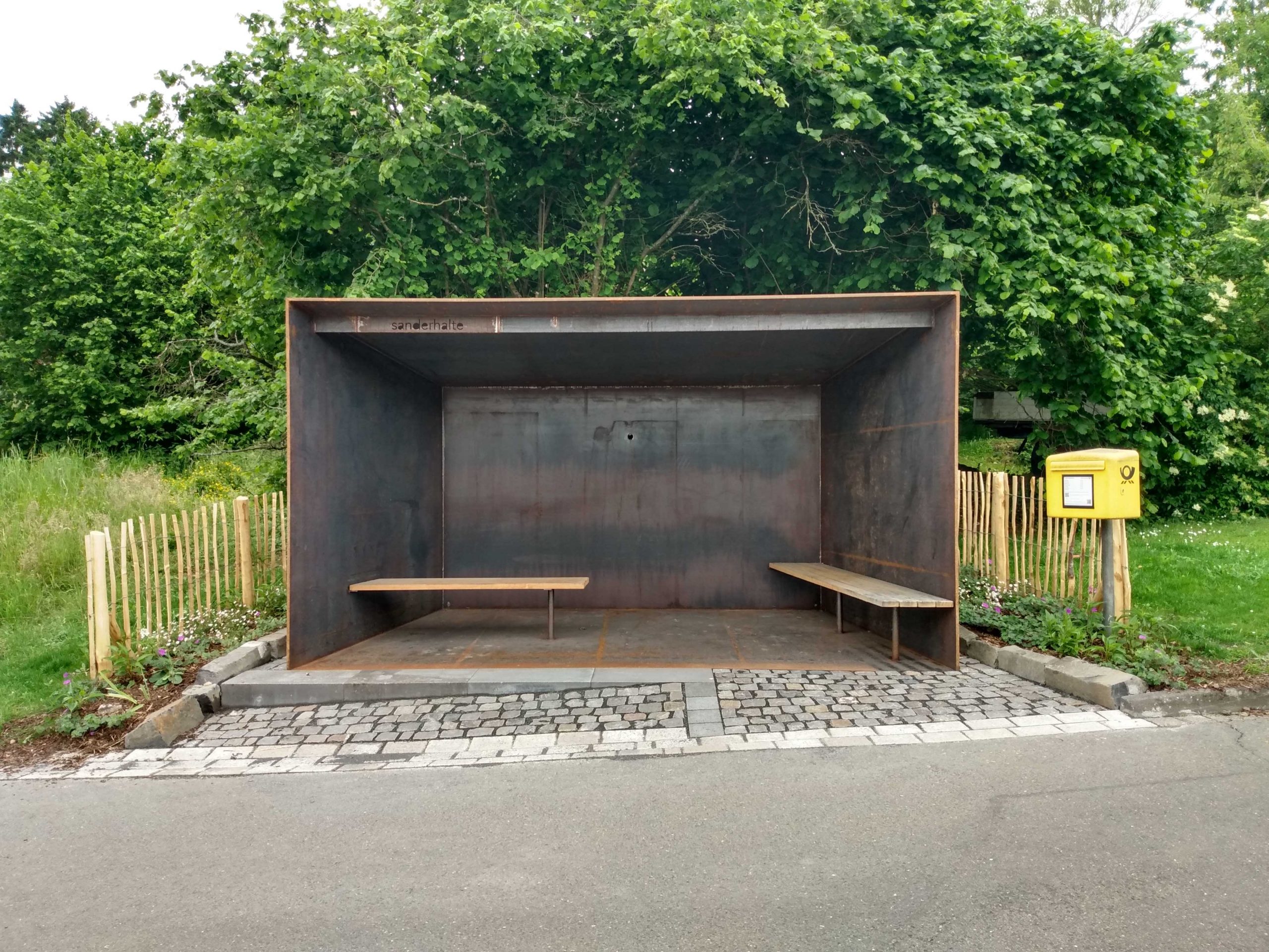 Haltestelle aus Stahl mit Holzbänken, davor abschüssige Straße